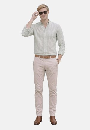 Joven ajustándose las gafas de sol, vestido con una camisa de rayas abotonada, pantalones beige, cinturón marrón, reloj y botas marrones, de pie contra un fondo liso.