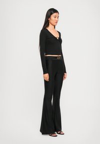 Black long-sleeve top with a twisted front detail and circular hardware; paired with black flared pants and strappy black sandals.