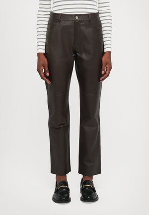 Persoon in donkerbruine leren recht-pijpen broek, zwarte loafers met gouden kettingen, en een witte long-sleeve shirt met zwarte strepen.
