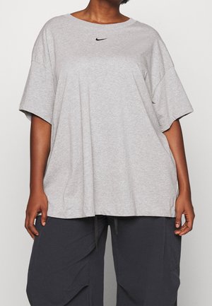 Persoon draagt een losvallend lichtgrijs T-shirt met korte mouwen, voorzien van een klein zwart Nike-logo, en donkergrijze broek tegen een effen achtergrond.