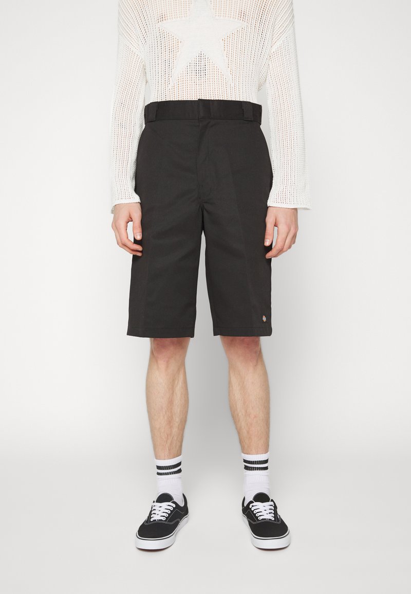 Black shorts with a straight cut, belt loops, side pockets, and a small logo detail near the hem. Worn with black sneakers and striped socks.