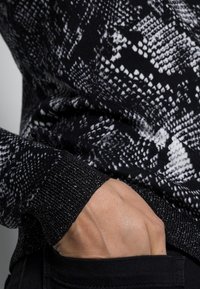 Svartvit tröja med mönster av ormar, med strukturerad stickning, glittrande svarta muddar och en sidoficka.