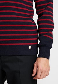 Granatowy sweter z czerwonymi poziomymi paskami, faktura ribbed oraz małym haftowanym logo kotwicy na dolnym brzegu. Długie rękawy i dopasowany krój.