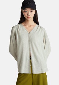 Camicia button-up a righe crema con scollo a V, maniche lunghe e vestibilità rilassata, abbinata a pantaloni verde oliva. Il cappellino presenta un logo nero.