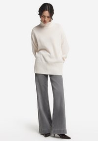 Weißer Pullover mit hohem, geripptem Kragen und geripptem Saum, kombiniert mit weitem grauen Jeans. Glatte Textur, neutrale Farben.