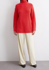 Femme portant un pull ample à col roulé rouge vif, un pantalon large crème et des chaussures noires à bout pointu sur fond clair.