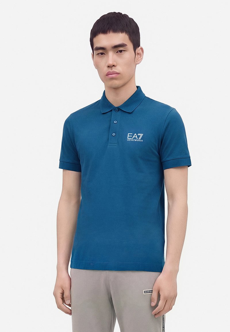 Jeune homme portant un polo bleu EA7 Emporio Armani et un pantalon gris clair avec un écusson logo EA7 sur la partie supérieure de la cuisse.