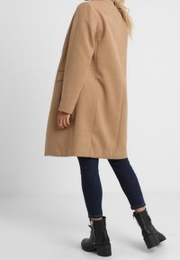 Cappotto in misto lana color cammello con taglio dritto, due tasche laterali e una cucitura pulita sul retro, abbinato a jeans scuri e stivali neri con zip alla caviglia.