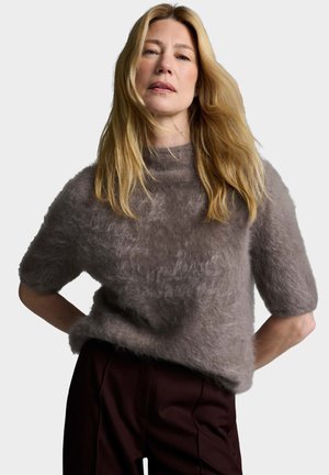 Frau mit langen blonden Haaren trägt einen flauschigen grauen kurzärmeligen Pullover und dunkle Hosen, steht mit den Händen in der Hüfte vor einem schlichten Hintergrund.
