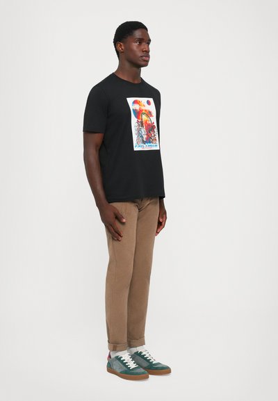 Black short-sleeve t-shirt with colorful graphic print, paired with tan trousers and green sneakers with white laces and gum soles.