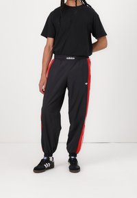 T-shirt nera, pantaloni della tuta neri a strisce rosse con cintura elastica e sneakers nere con accenti bianchi e blu.