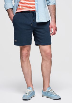 Mand iført marineblå shorts med snøre, en lys denimskjorte over en orange stribet skjorte og lyseblå sneakers, står med hænderne i lommerne.