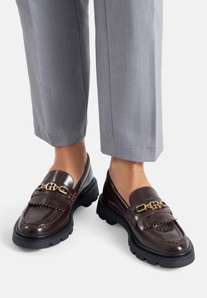 Braune Leder-Loafers mit Fransenakzent, goldfarbenen Beschlägen und strukturierter Plateau-Sohle, kombiniert mit grauen Hosen.