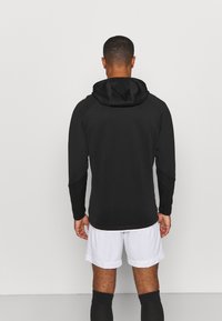 Zwarte hoody met een gestructureerde afwerking, lange mouwen en een ronde zoom, gedragen over witte shorts en zwarte voetbalsokken.