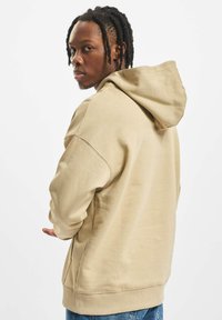 Beige hooded sweatshirt with a soft texture, featuring a relaxed fit and ribbed cuffs. Model is shown from the back, with hands gesturing.