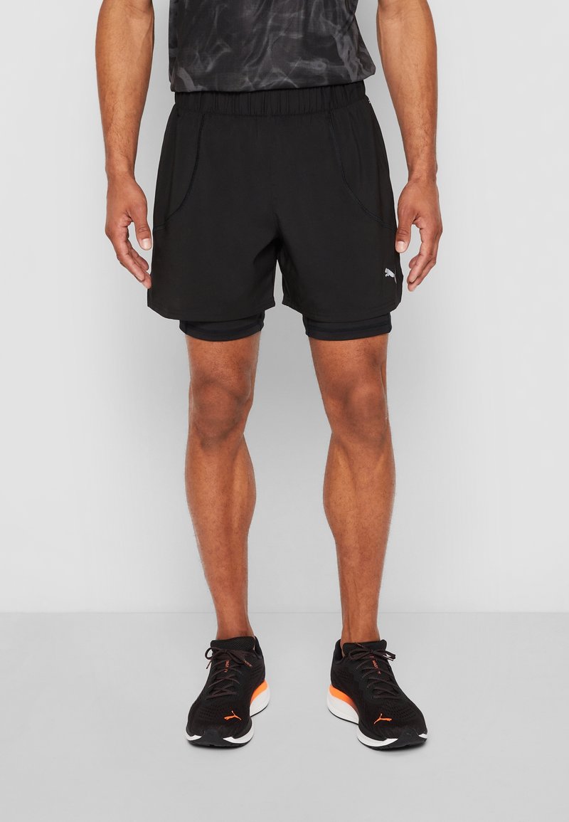 Mies, joka käyttää mustia kaksikerroksisia juoksushortseja ja mustia juoksukenkiä oransseilla yksityiskohdilla, seisoo tasaisen harmaan taustan edessä.