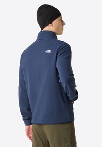 Giacca in pile blu navy con colletto alto, maniche lunghe, tasche laterali e un piccolo logo bianco sulla parte superiore della schiena. Il tessuto sembra morbido.