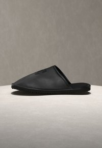 Μαύρο λαστιχένιο παντόφλα τύπου slip-on με υφή στην σόλα και διακριτικό ανάγλυφο λογότυπο στην κορυφή, απεικονίζεται σε πλαϊνή όψη πάνω σε φωτεινή επιφάνεια.