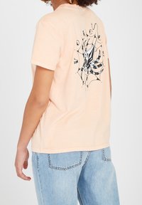 Perzikkleurig T-shirt met korte mouwen, voorzien van een zwart en teal spinnenprint op de achterkant, gemaakt van glad katoen.