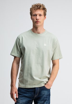 Lichtgroene katoenen T-shirt met korte mouwen, ronde hals en een klein logo op de borst. Draagt met donkerblauwe jeans.
