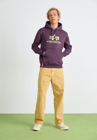 Sweatshirt à capuche violet avec un logo vert, poche frontale, et capuche avec cordon, associé à un pantalon beige et des baskets claires.