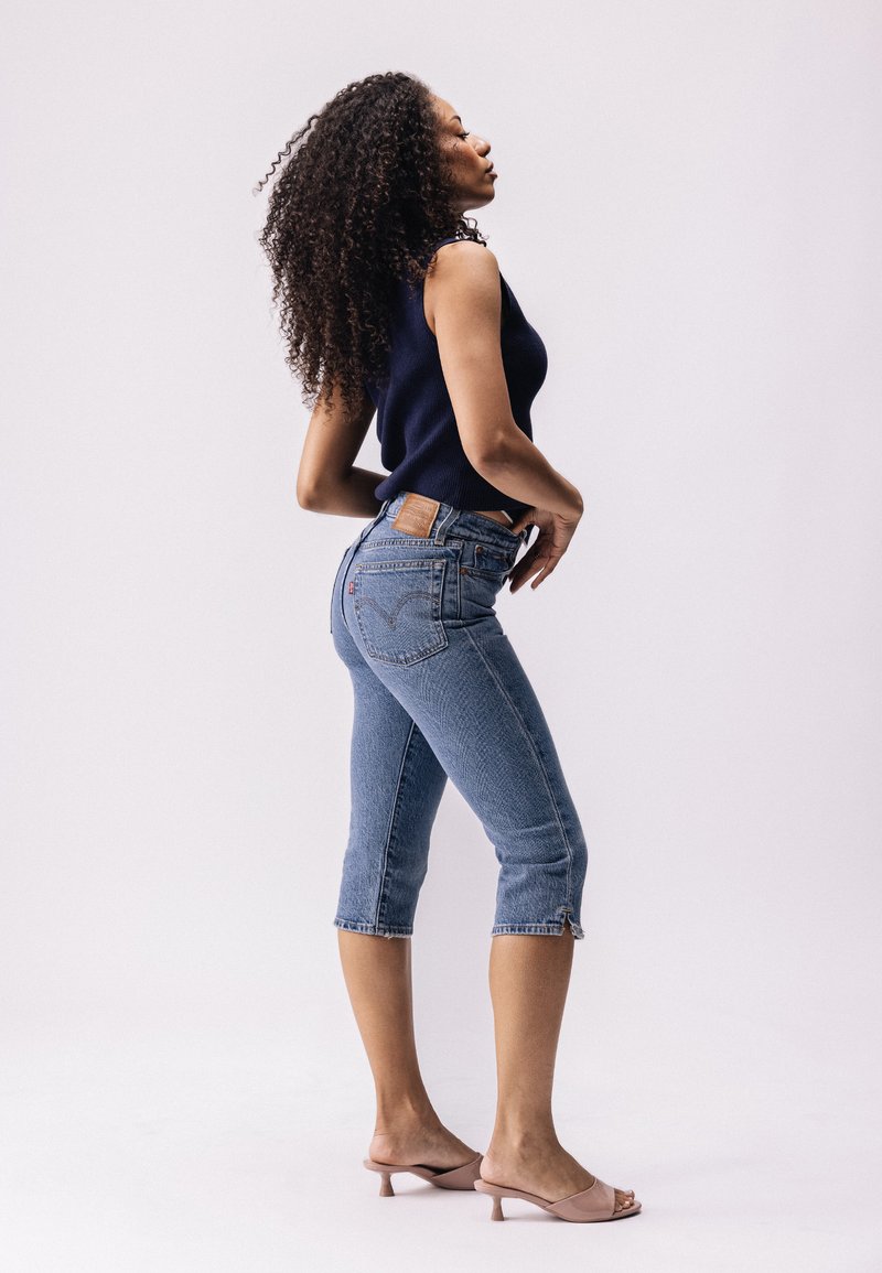 Haut sans manches bleu marine associé à un jean capri en denim bleu. Le jean présente des surpiqûres contrastées et un détail zippé de style cargo au niveau de l'ourlet. Talons légers.