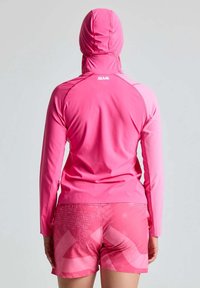 Langärmliger pinkfarbener Sport-Hoodie mit Kapuze, der eine taillierte Passform und kontrastierende Akzente aufweist. Kombiniert mit pinkfarbenen, gemusterten Shorts.