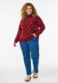 Maglione rosso a maglia con un motivo testurizzato, abbinato a jeans in denim blu e stivaletti beige alla caviglia. Il maglione ha un collo alto e orlo a costine.