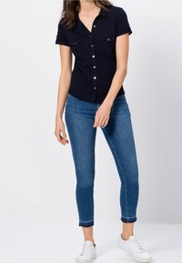Žena v černé krátké košili na knoflíky, modrých úzkých džínách a bílých teniskách stojící před jednobarevným pozadím.