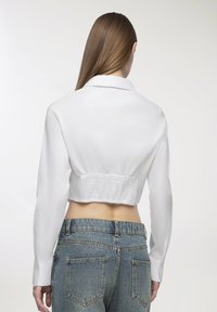 Camicetta bianca corta con maniche lunghe e colletto; presenta una schiena arricciata, abbinata a jeans in denim blu a vita alta.