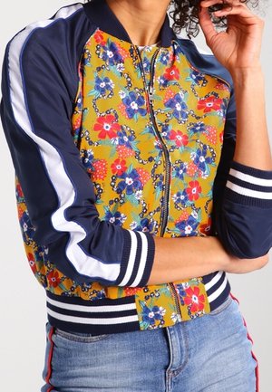 Personne portant une veste bomber fleurie et colorée avec des manches bleu marine et des rayures blanches, associée à un jean bleu clair.