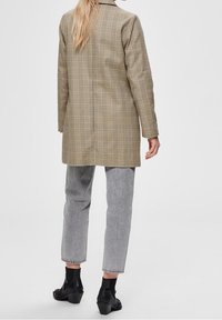 Kvinde med langt blonde hår iført en beige ternet blazer, grå cropped jeans og sorte højhælede ankelstøvler, står mod en ensfarvet grå baggrund.