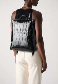 Mochila de charol negra con textura de cocodrilo, que cuenta con un cierre de cremallera y un logo en relieve. Las correas ajustables completan el diseño.