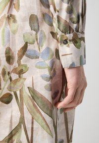 Rochie cu model floral, realizată dintr-o țesătură ușoară și transparentă. Prezintă frunze verzi și albastre moi, manșete cu nasturi și un design curgător, texturat.