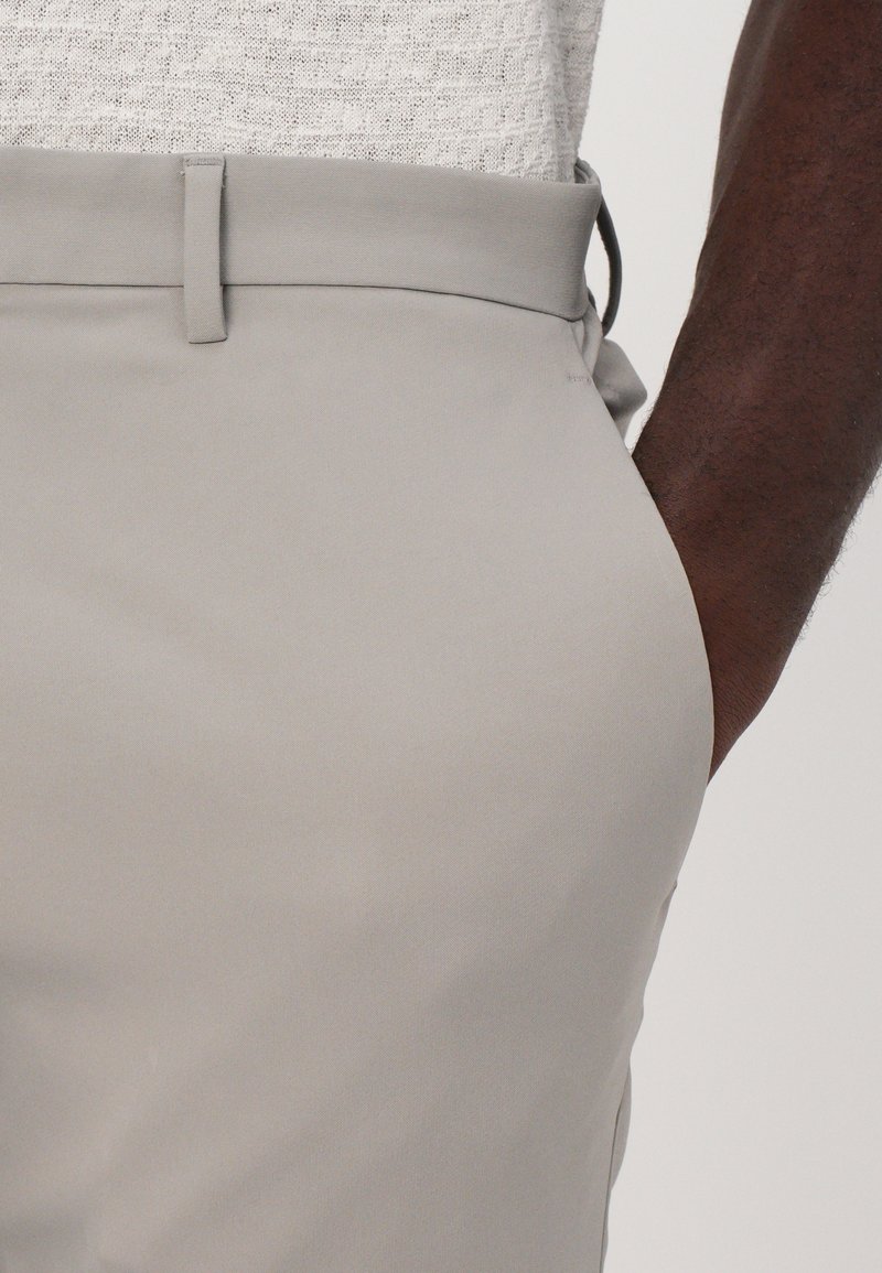 Pantalons gris clair avec une texture lisse et une coupe ajustée. Présente une taille plate, des passants de ceinture et un détail subtil à la taille.