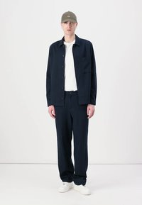 Navy blue shirt with buttons, white t-shirt underneath, navy pants, and white sneakers. Olive green cap. Plain background.