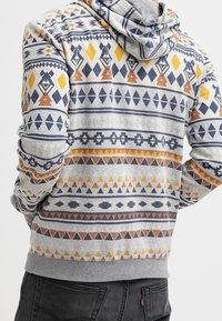 Sudadera con capucha de base gris, con patrones geométricos multicolores en amarillo, naranja y azul marino. Tejido texturizado, capucha visible.