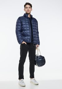 Navy quilted puffer jacket with high collar, zip pockets, worn over a dark top. Paired with black pants and white sneakers, holding a navy bag.