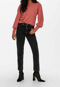 Žena v růžovém dlouhém svetru s detaily ve tvaru oka, černých vysokých džínách a černých síťovaných kotníkových botách na podpatku, stojící se rukou v kapse.