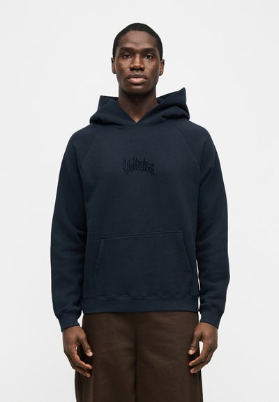 Άνδρας που φοράει μπλε σκουρόχρωμο φούτερ με κουκούλα και μπροστινή τσέπη, με μαύρο στυλιζαρισμένο κείμενο στο στήθος, συνδυασμένο με σκούρο καφέ παντελόνι, στέκεται μπροστά σε λευκό φόντο.