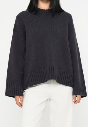 Femme portant un pull en maille gris foncé ample à larges manches, associé à un pantalon blanc, torse et bras visibles sur fond blanc.