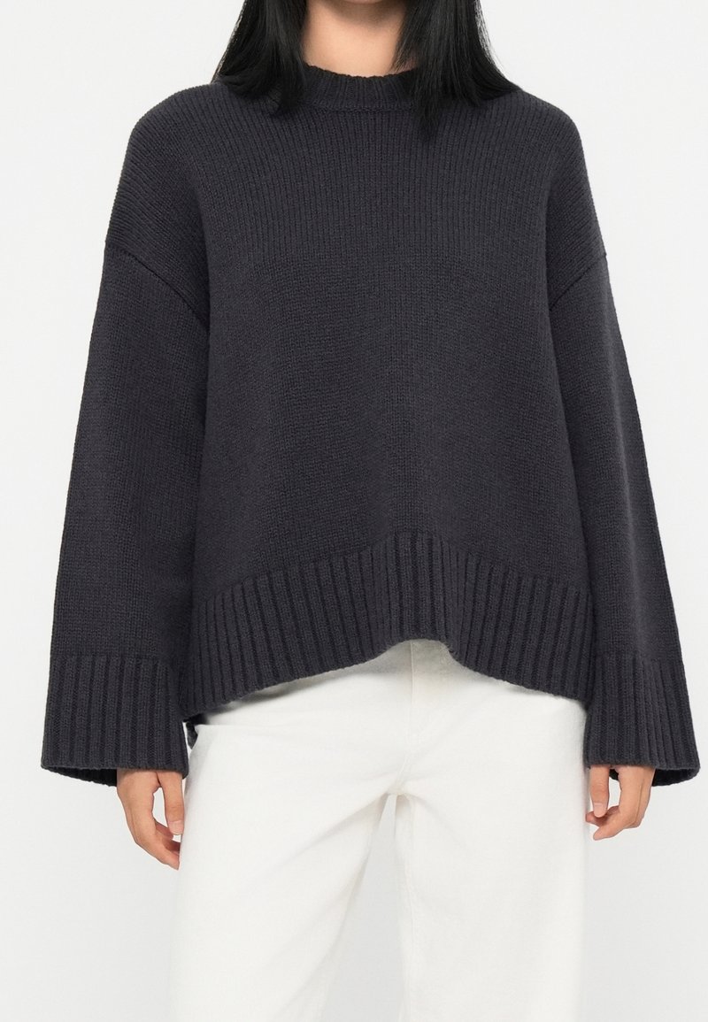 Femme portant un pull en maille gris foncé ample à larges manches, associé à un pantalon blanc, torse et bras visibles sur fond blanc.