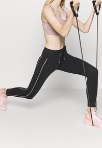 Černé sportovní legíny s růžovými bočními pruhy, elastickým pasem a kapsami, v kombinaci s růžovým sportovním podprsenkou a odpovídajícími teniskami.