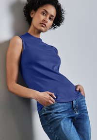 Ärm-lös blå topp med hög halsringning, gjord av ett slätt material. Bärs med blå denimjeans som framhäver en åtsittande silhuett och rena linjer.