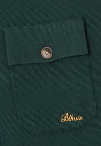 Mørkegrønn strikket stoff med en lommeflap med knapper og gullbrodert "Blktis"-tekst nederst til høyre på lommen.