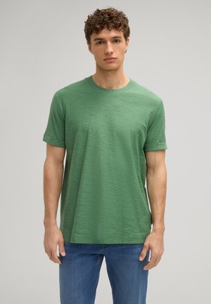 Grünes T-Shirt mit kurzen Ärmeln aus strukturiertem Stoff. Es hat einen Rundhalsausschnitt und aufgerollte Ärmel und wird mit blauen Jeans kombiniert.