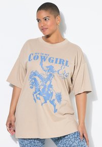 Beige oversize tričko s modrou grafikou kovboje na koni a textem "já v mé KOVBOJKOVÉ ÉŘE." Měkká bavlněná látka.