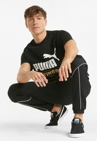 Černé tričko s bílým logem Puma a zlatým textem "KING", v kombinaci s černými tepláky s bílými bočními pruhy a černými teniskami.