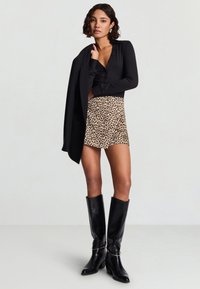 Schwarze Bluse mit langen Ärmeln, bedruckter Leoparden-Minirock und kniehohe schwarze Lederstiefel mit Absatz und metallic Akzent. Modell posiert selbstbewusst.