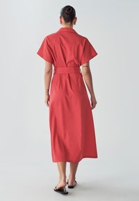 Czerwona, średniej długości sukienka z krótkimi rękawami, z paskiem w talii i kołnierzykiem. Gładki materiał o luźnym kroju. Na nogach sandały.